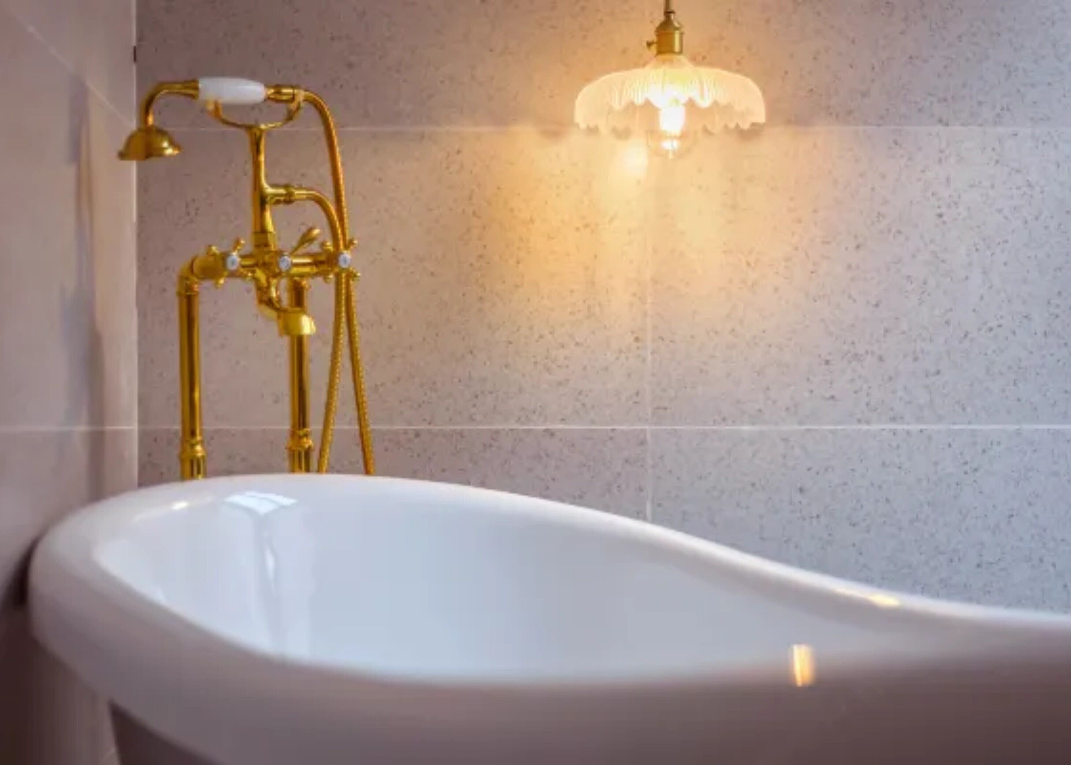 A bathroom with a freestanding bathtub, a gold-colored showerhead, and a wall-mounted light fixture.