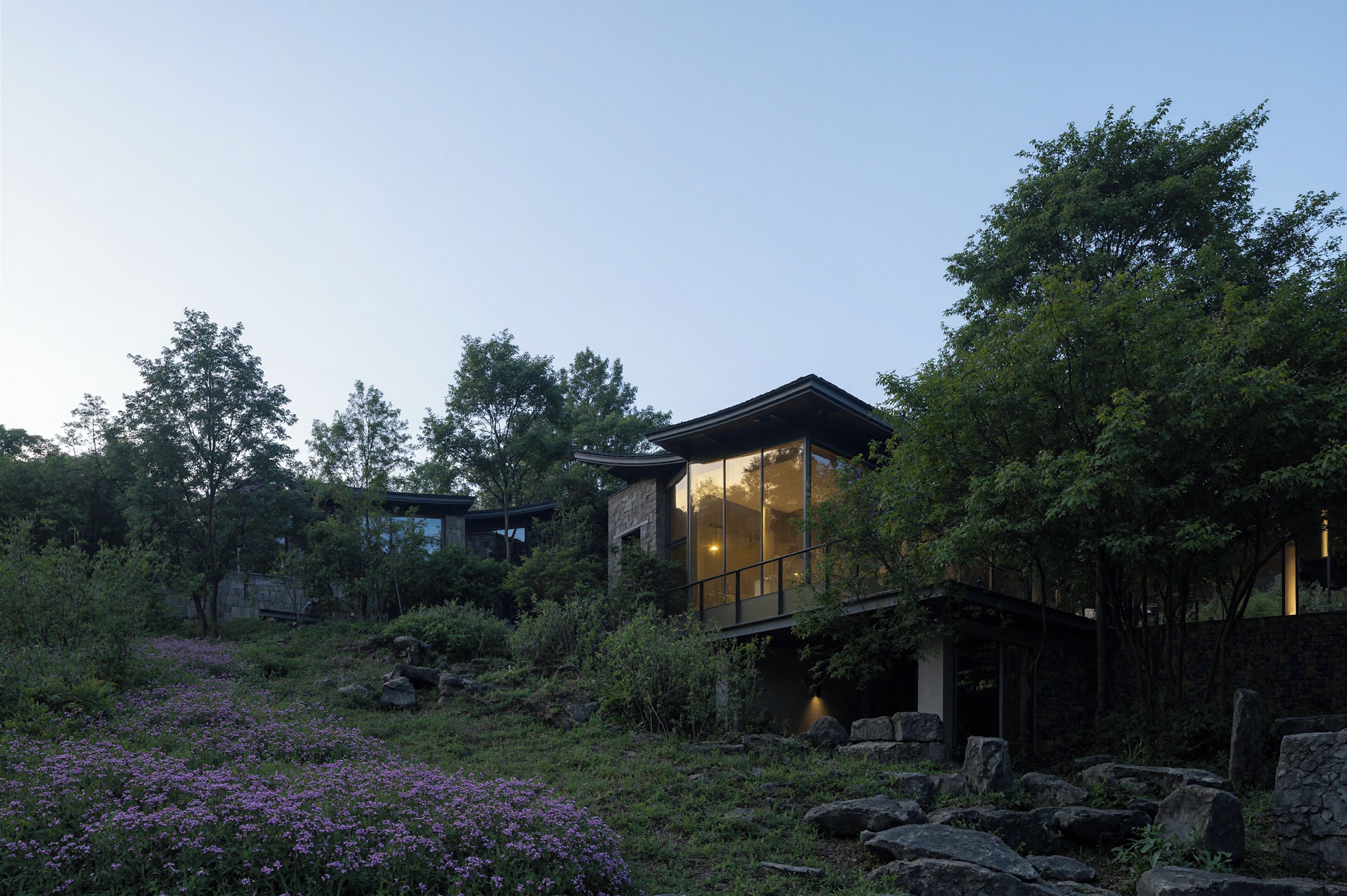 Ein modernes Haus mit Glasfassade, umgeben von üppigem Grün und violetten Blumen.