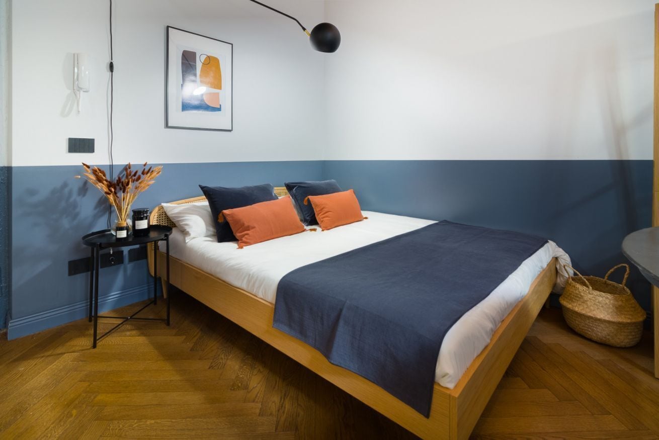 A bedroom with a bed, a bedside table, a lamp, a wall-mounted phone, and a framed picture.
