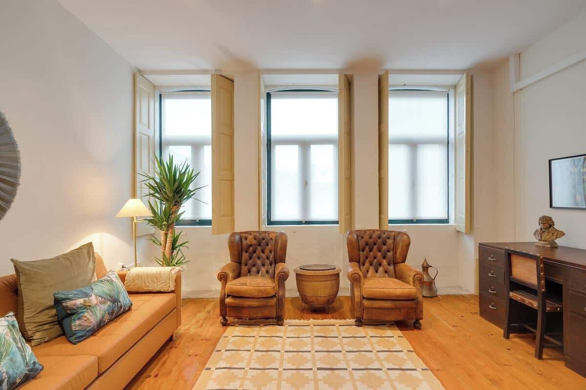A living room with a sofa, two armchairs, a coffee table, and a desk with drawers. Large windows with blinds are on the wall.