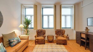 Olohuoneessa on sohva, kaksi nojatuolia, sohvapöytä ja työpöytä, jossa on laatikoita. Suuret ikkunat, joissa on sälekaihtimet, ovat seinällä.