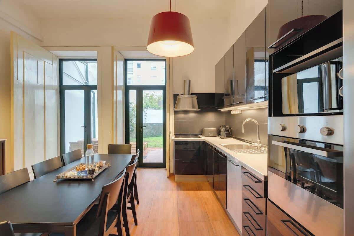 A modern kitchen with a dining area, featuring a large window, stainless steel appliances, and a red pendant light.