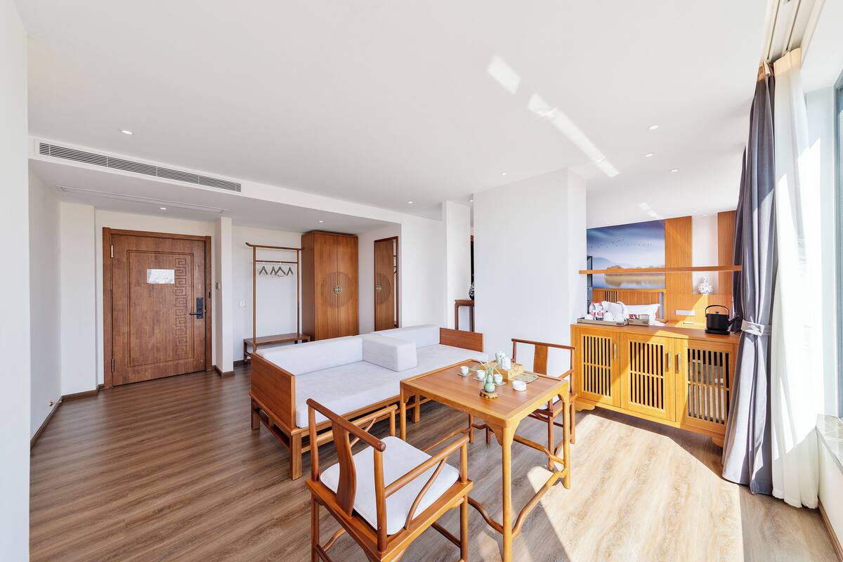 A modern hotel room with a wooden floor, a white sofa, a dining table with chairs, a kitchen area, and a large window with curtains.