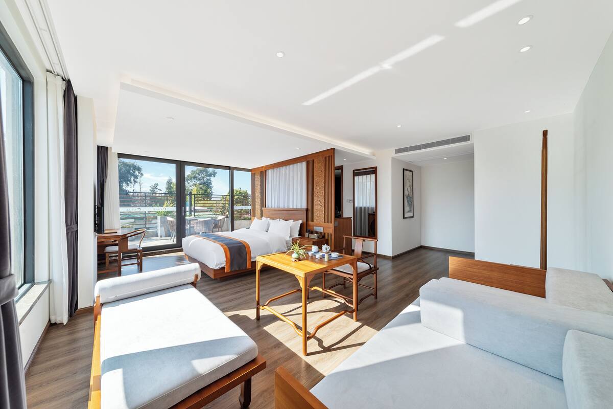 A modern hotel room with a large bed, a sofa, a dining table, and a balcony with a view.