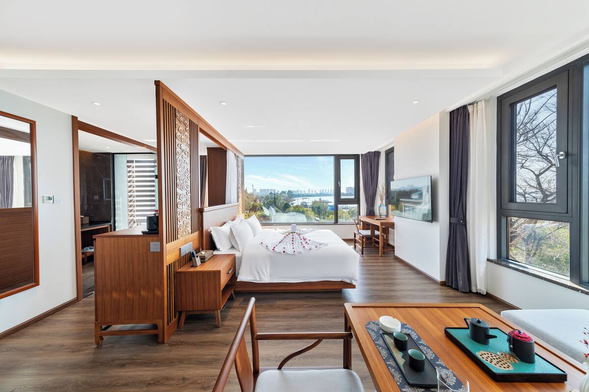 A modern hotel room with a large bed, wooden furniture, a dining area, and a view of the city.