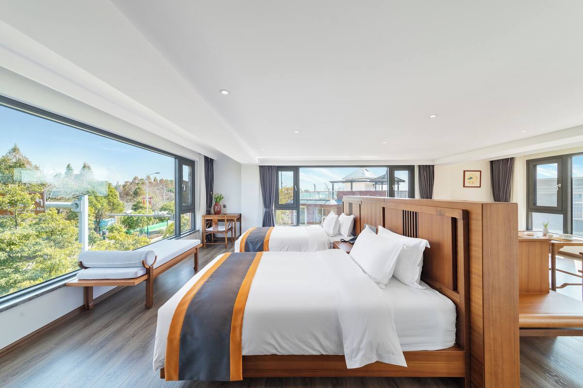 A hotel room with two beds, a wooden headboard, a bench, a desk, and a large window offering a view of trees and buildings.