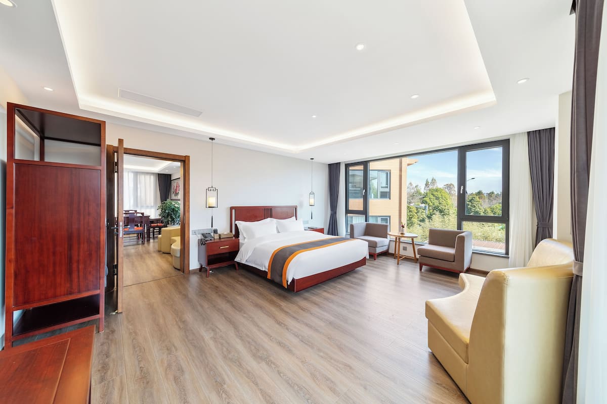 A modern hotel room with a large bed, a seating area with a sofa and armchairs, a wooden floor, and large windows offering a view of the outdoors.