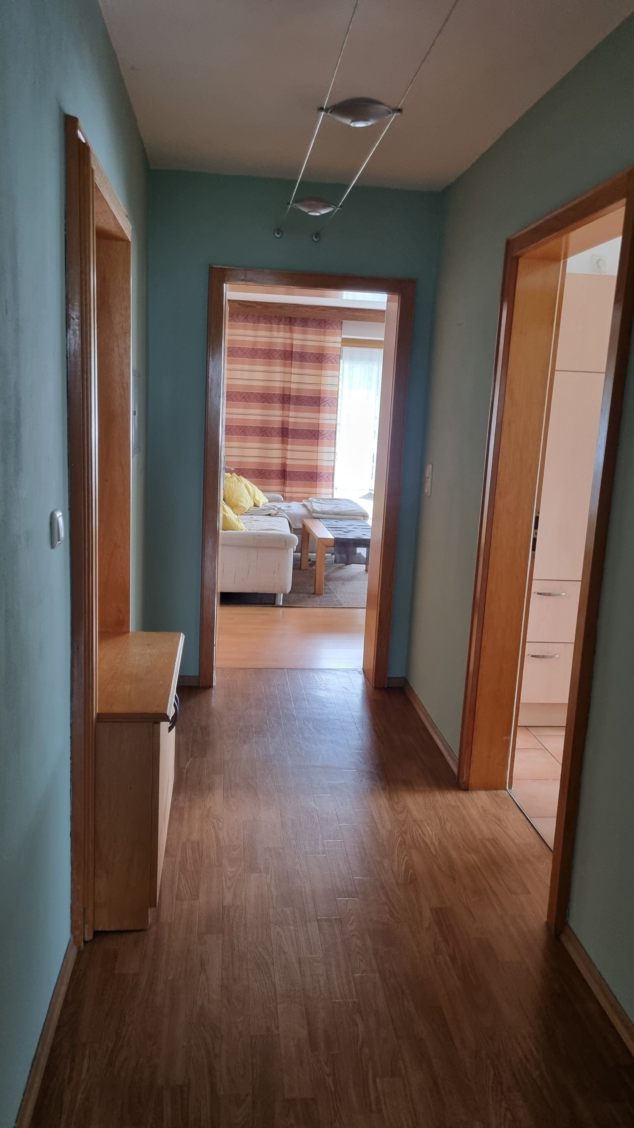 Un couloir mène à une chambre qui contient un lit, une table de chevet et une fenêtre avec des rideaux.