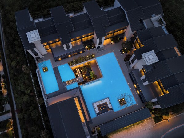 An aerial view of a modern hotel with multiple swimming pools, a rooftop garden, and a well-lit outdoor area.