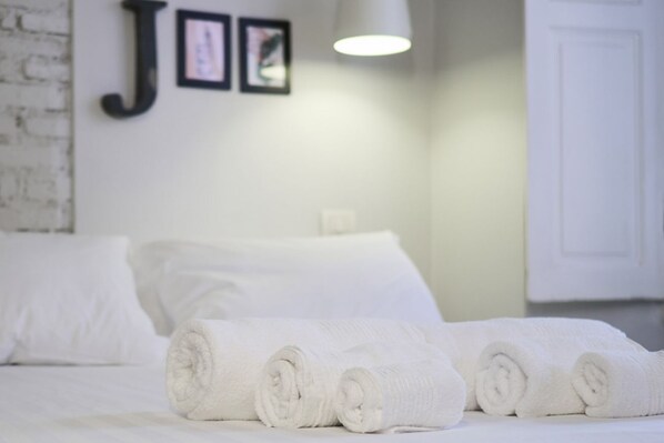 A neatly made bed with rolled white towels, a white wall with a framed letter 'J', and a white door.