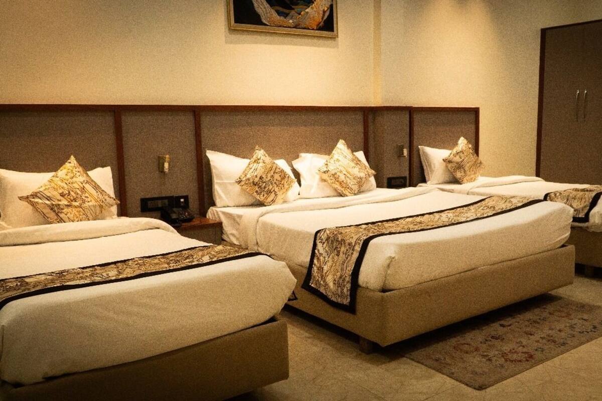 A hotel room with three beds, each with a patterned bedspread and matching pillows. There is a wooden headboard, a nightstand with a lamp, and a wardrobe.