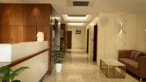 Ein Hotel-Empfangsbereich mit einer Rezeption, einer Zimmerpflanze, einer Couch und einem Gang zu weiteren Zimmern.