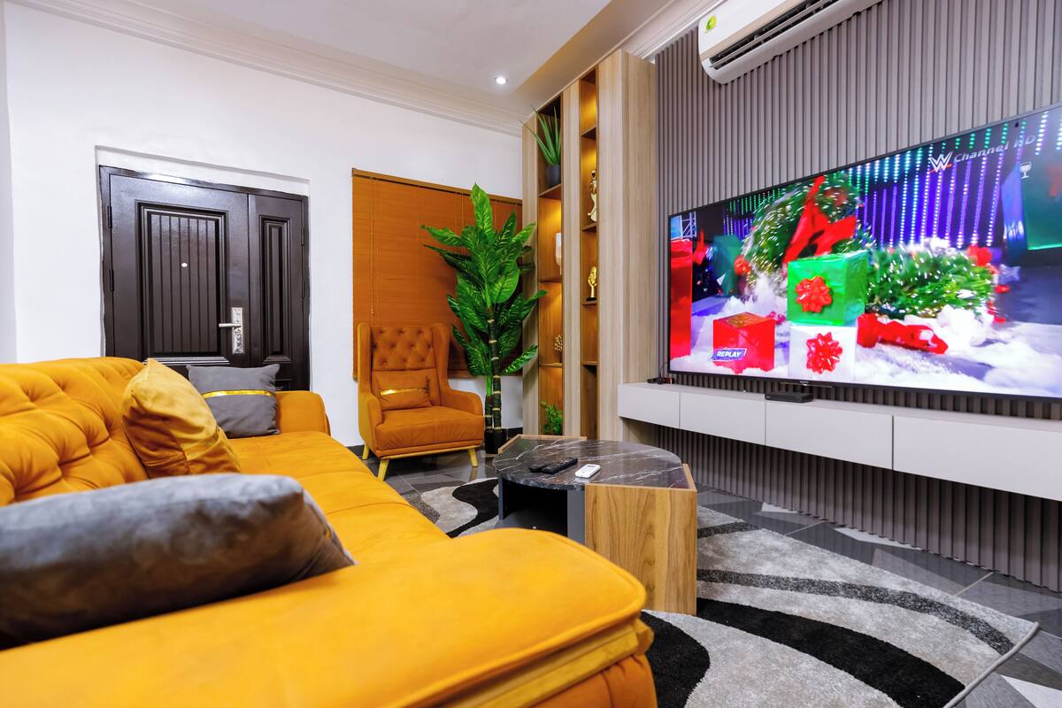 A modern living room with a large flat-screen TV, a comfortable orange sofa, a wooden coffee table, and a potted plant.