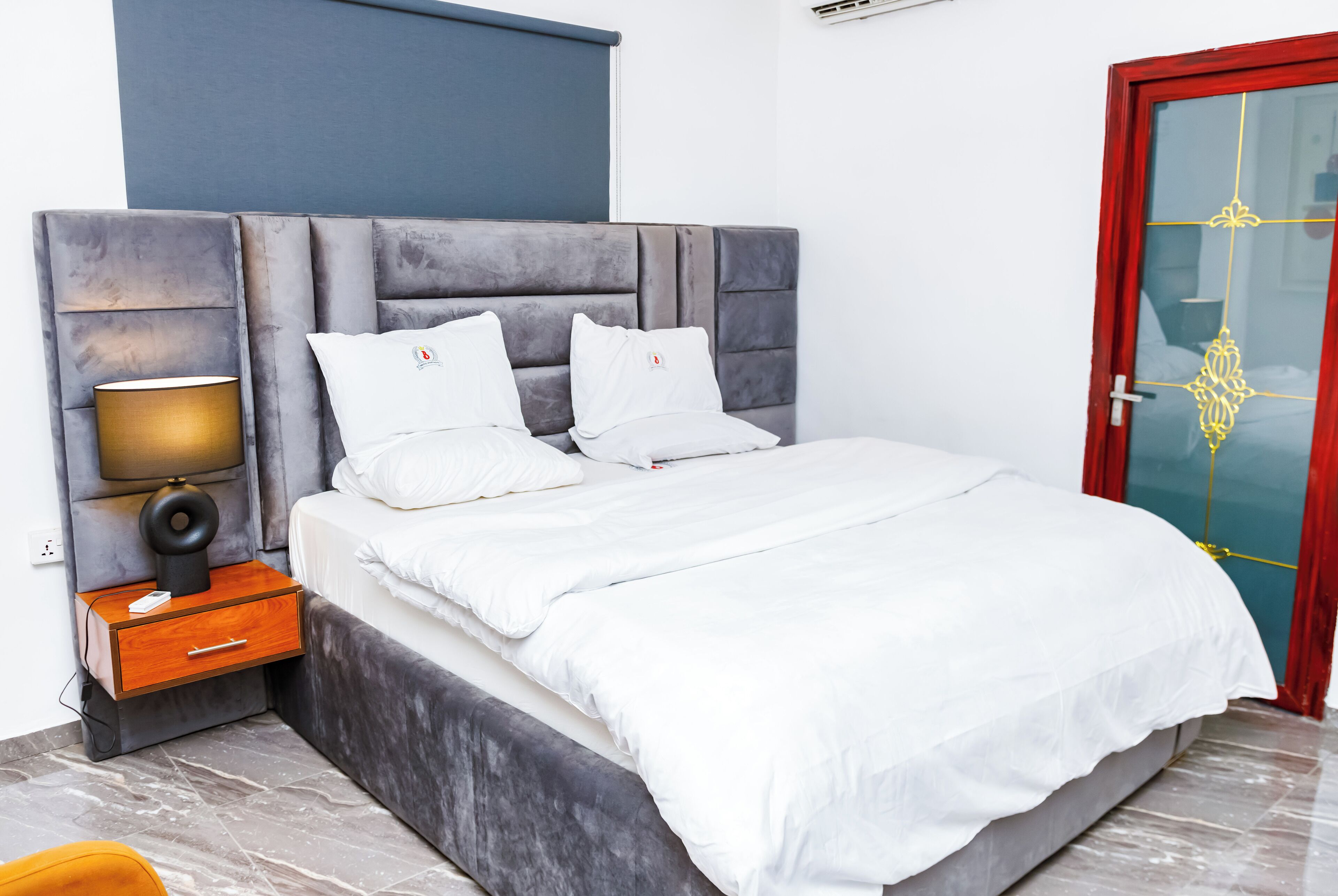 A modern bedroom with a large bed, a nightstand with a lamp, and a red-framed door.