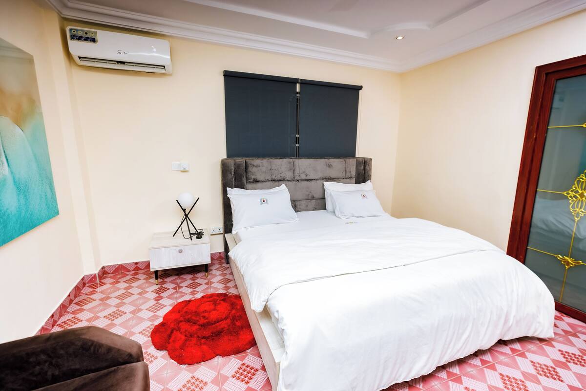 A bedroom with a bed, a nightstand, a red pouf, and a window with blinds.