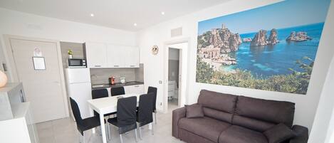 A modern living room with a brown sofa, a dining table with chairs, a kitchen area, and a large scenic painting on the wall.