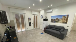A spacious living area with a grey sofa, a kitchen area with a black countertop, and a large window with blinds. - City Life Palermo (Palermo)