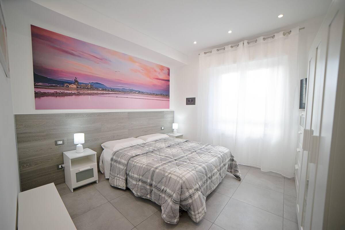 A bedroom with a bed, nightstands, a wall-mounted lamp, a painting, and a window with curtains.