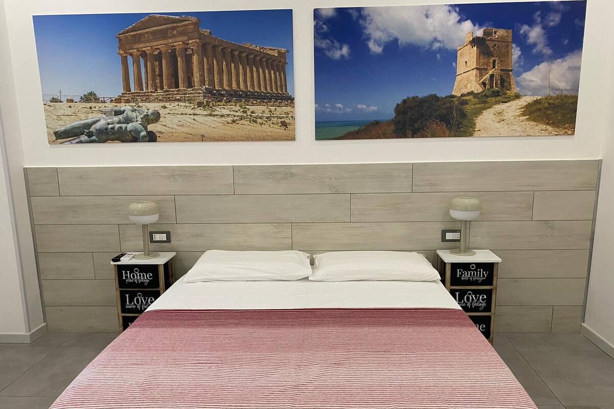 A bedroom with a bed, two nightstands, and two wall-mounted lamps. There are two framed pictures above the bed.