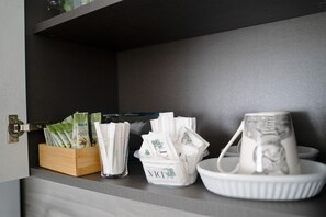 Eine Küchenarbeitsplatte mit einer Holzkiste voller Teebeuteln, einem klaren Glasbehälter ebenfalls mit Teebeuteln und einer weißen Keramiktasse auf einem weißen Teller.