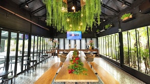 A spacious indoor area with a long wooden table decorated with greenery and flowers, surrounded by chairs and tables.