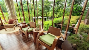A room with a wooden floor, two wooden chairs with green cushions, a wicker chair, a small round table, and a large window with a view of greenery.
