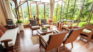 A spacious room with wooden flooring, a large window offering a view of lush greenery, and a seating area with multiple chairs and a sofa.