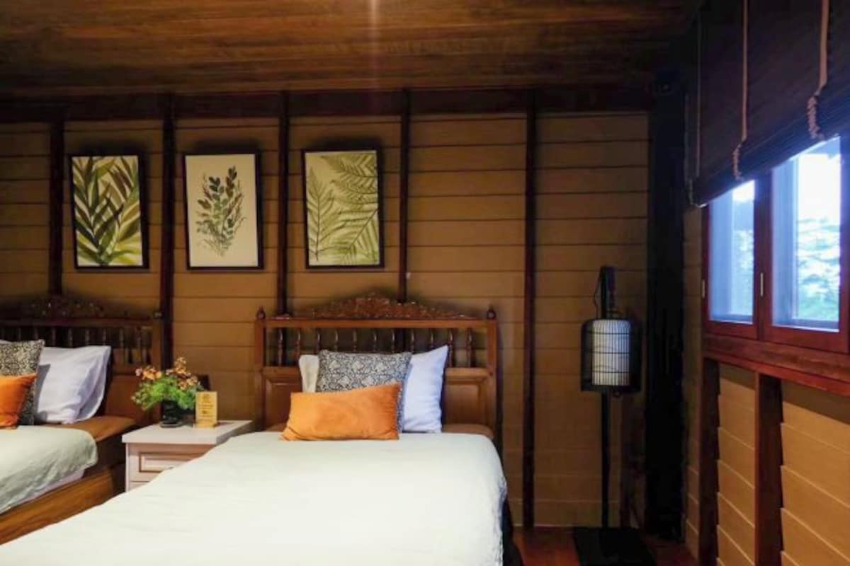 A wooden cabin room with two beds, a nightstand with a lamp, and framed botanical prints on the wall.