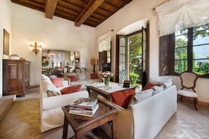 Ein geräumiges Wohnzimmer mit Holzbalkendecke, weißen Sofas, einem Holztisch mit Büchern und Blick ins Grüne durch große Fenster.