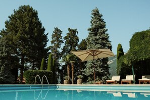 Une piscine entourée de chaises longues et d’un parasol, bordée de grands arbres et avec une montagne en toile de fond.