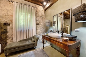 Une salle de bain rustique avec un mur en pierre, un meuble-lavabo en bois, un grand miroir et un banc rembourré.