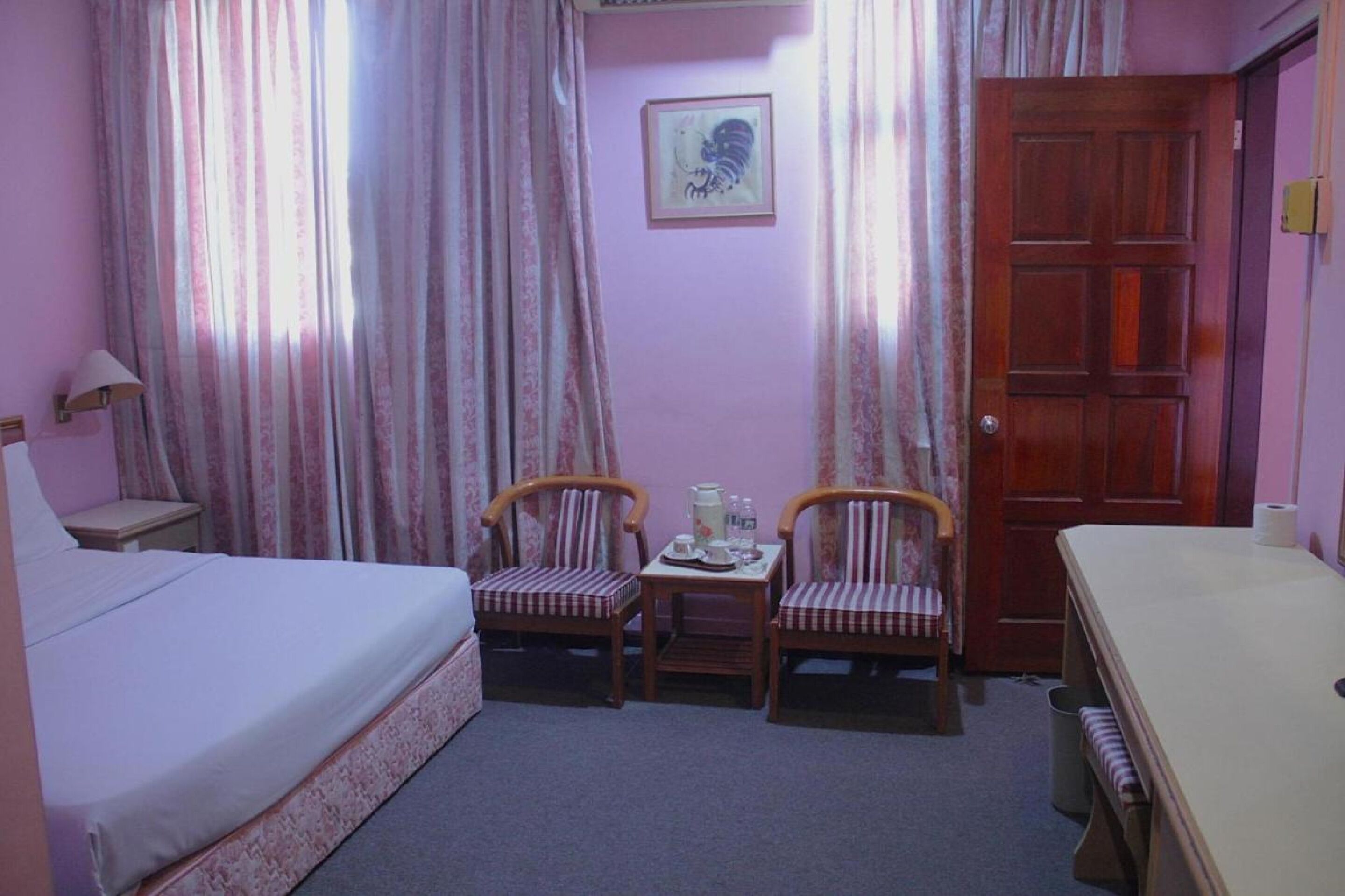 A small, cozy room with a bed, two chairs, a small table with a tea set, and a wooden door.