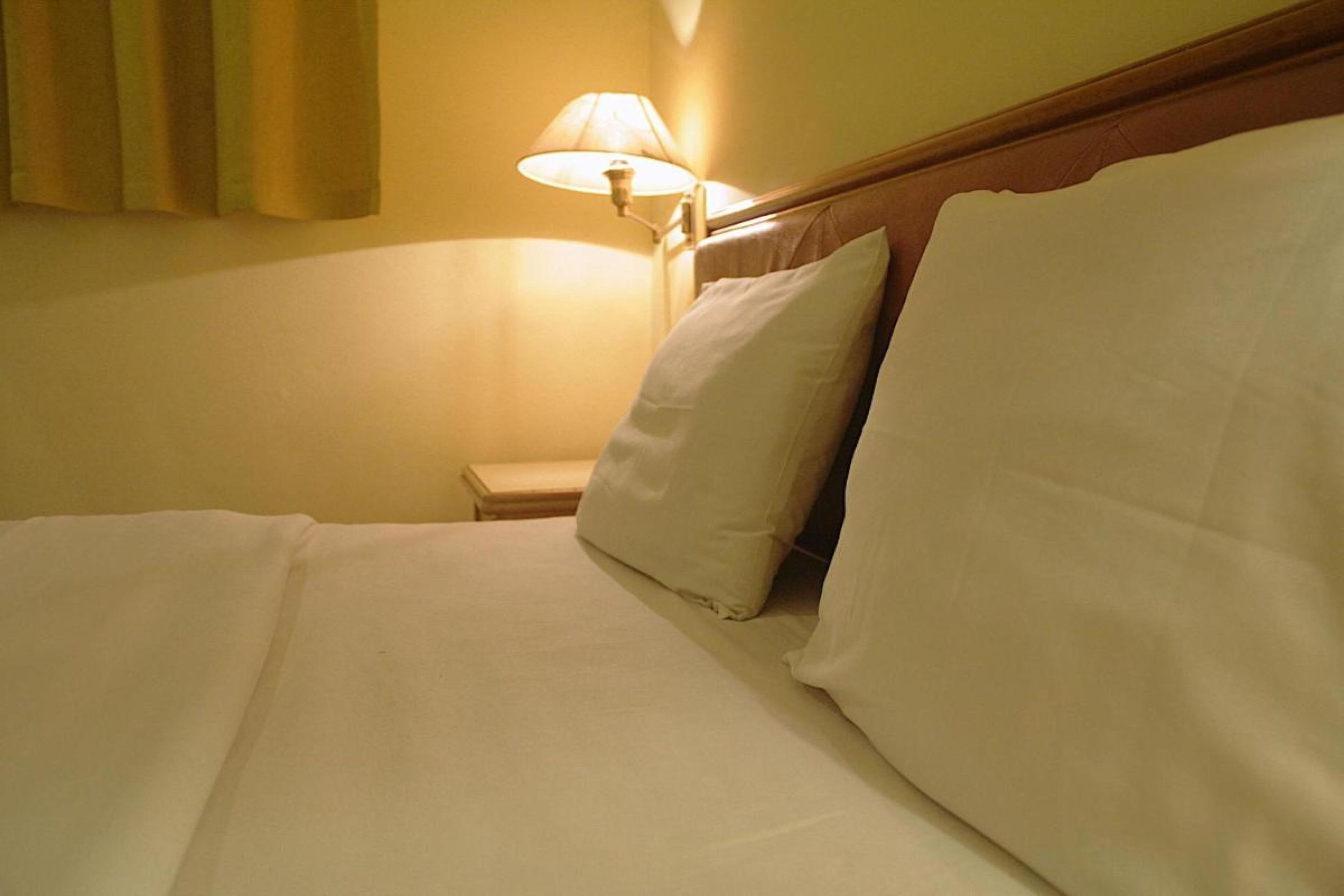 A neatly made bed with white linens and pillows, a bedside lamp, and a wooden headboard.