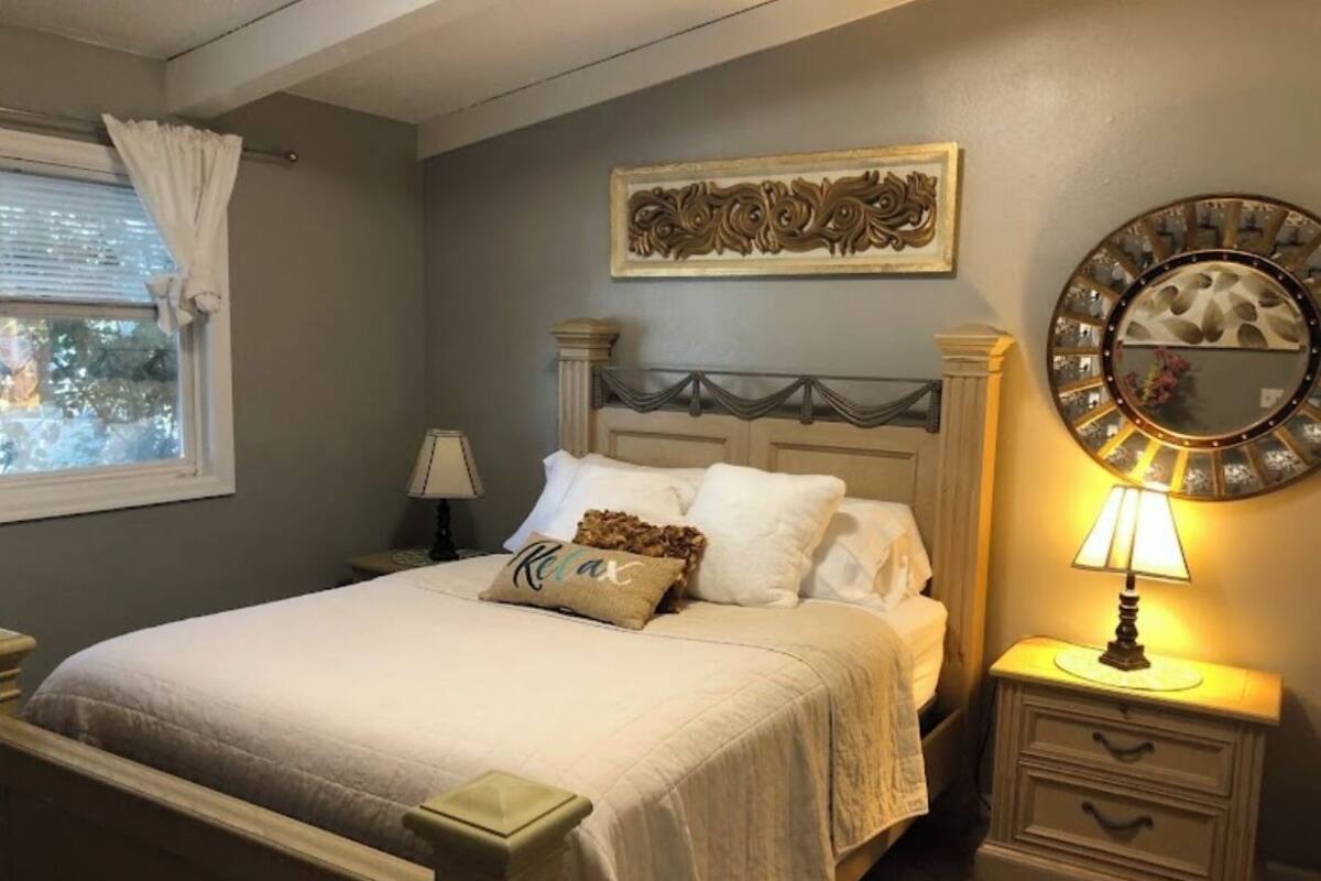 A bedroom with a bed, nightstands, a lamp, a decorative mirror, and a window with curtains.