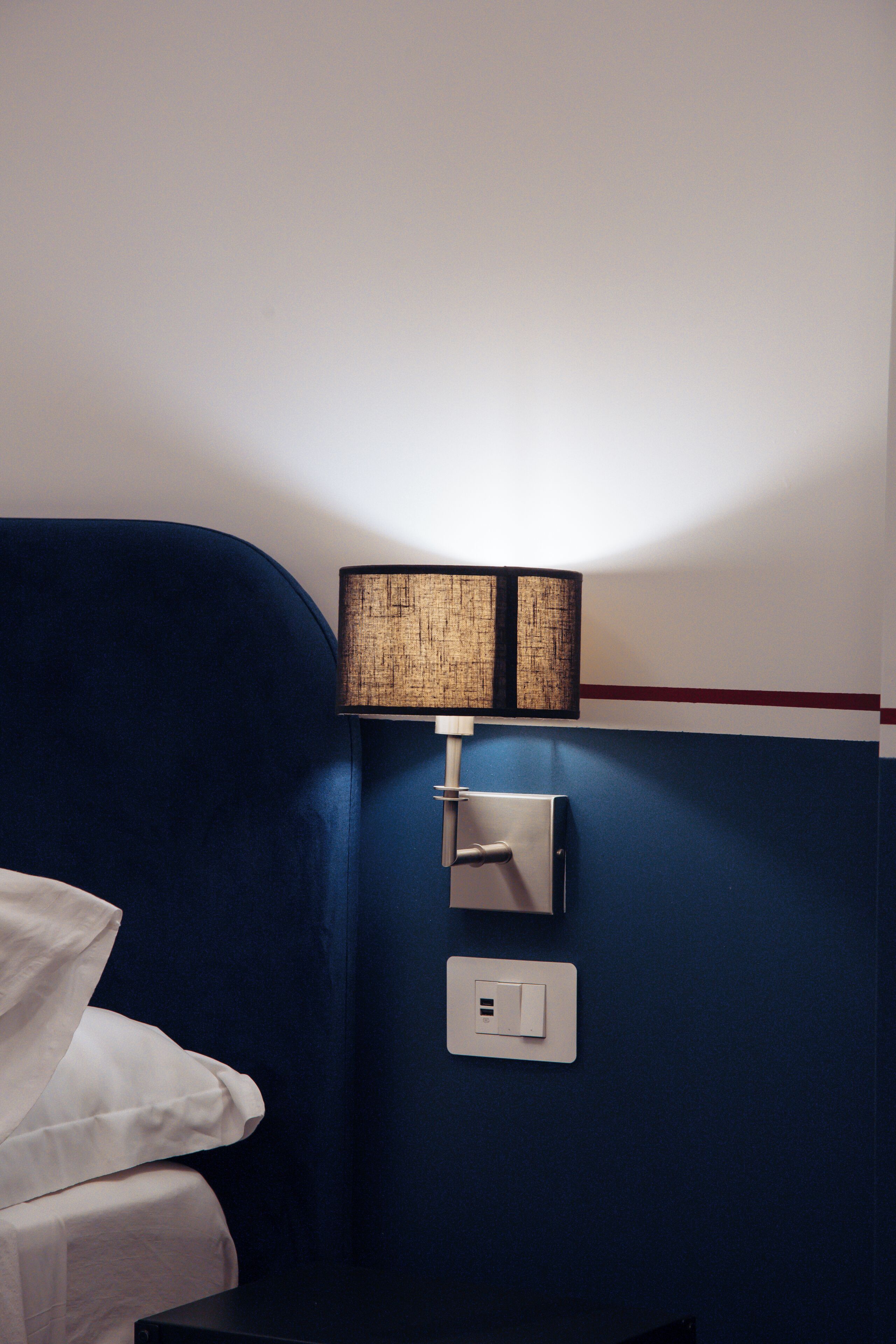 Habitación de hotel con pared azul, cama con ropa blanca, lámpara de pared y toma de corriente.
