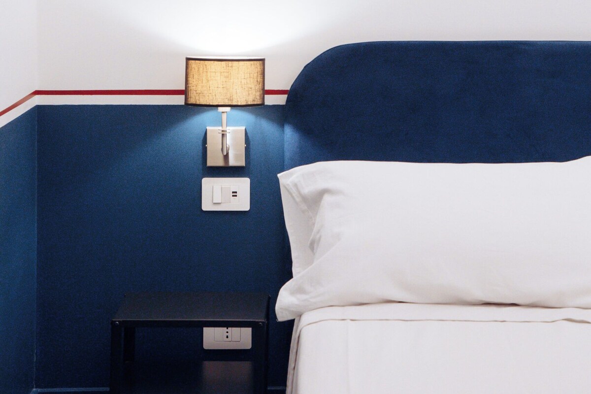 A hotel room with a bed, a nightstand, a lamp, and a wall socket.