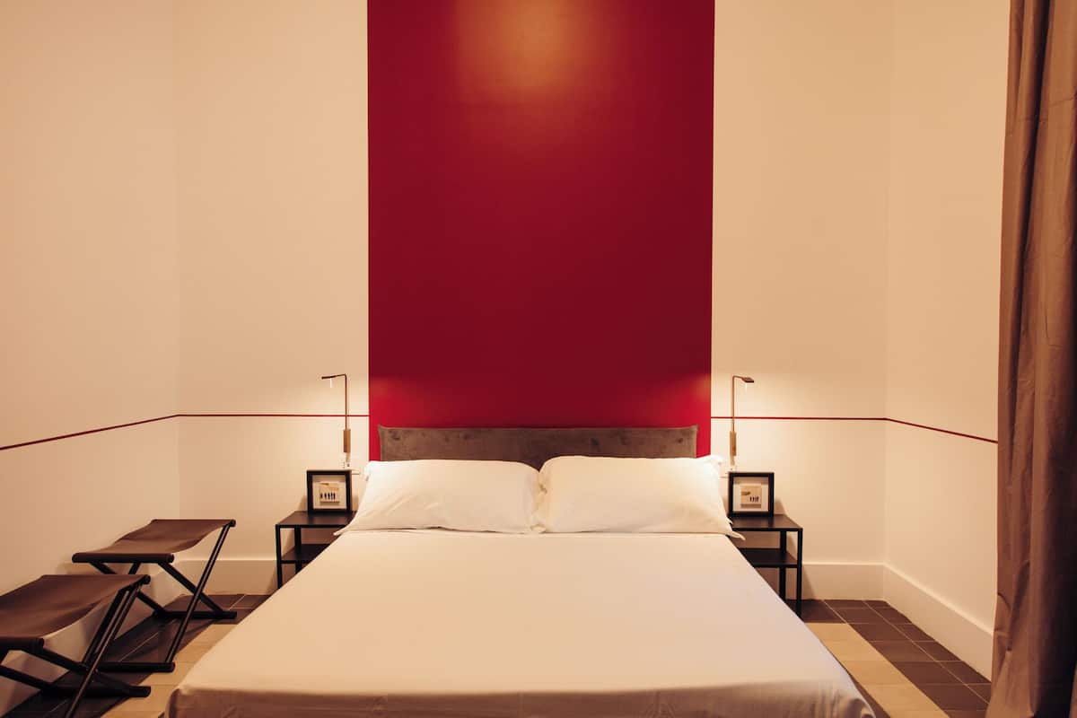 A modern hotel room with a large bed, two bedside tables, a nightstand, a chair, and a red accent wall.