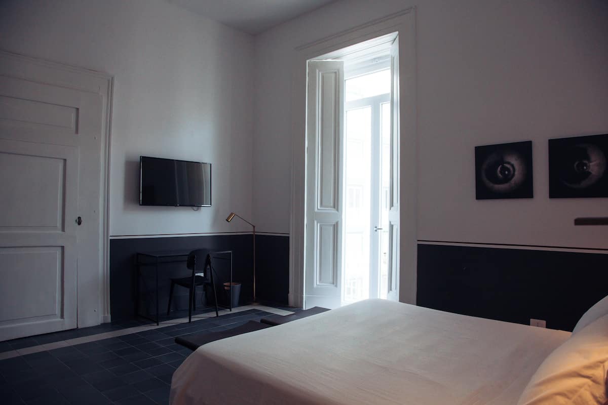 A hotel room with a bed, a desk with a chair, a television on the wall, and two framed pictures.