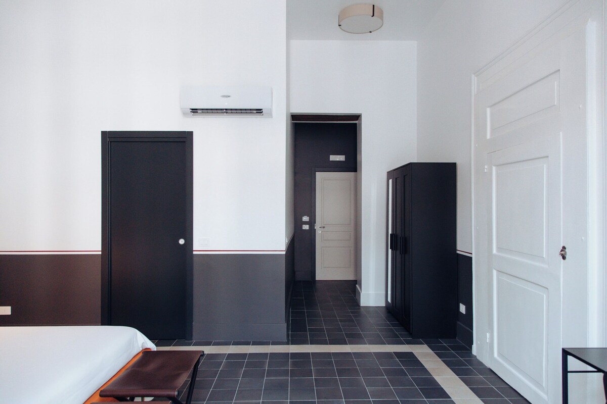 Une chambre d’hôtel comprenant un lit, un banc, une armoire et une porte donnant sur un couloir.