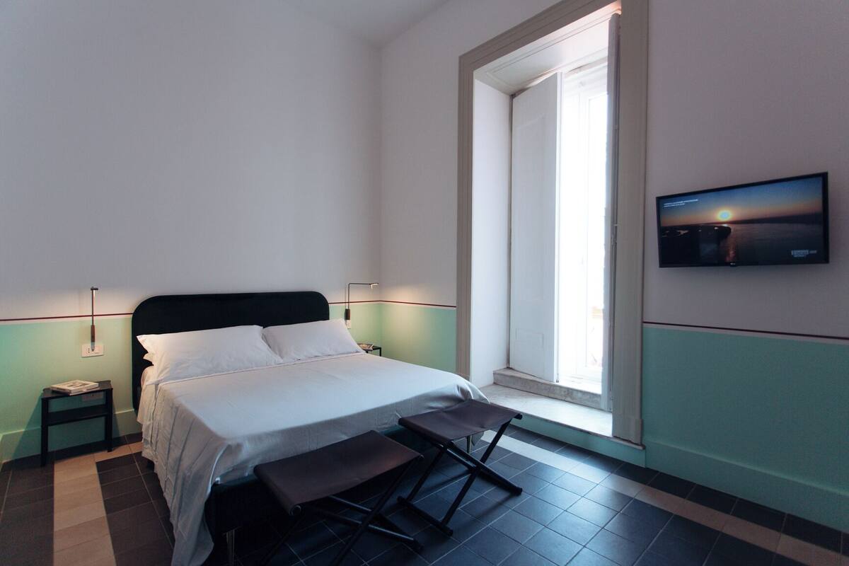 A neatly made bed with white linens, a bedside table with a lamp, a bench at the foot of the bed, and a large window with a view of the sunset.