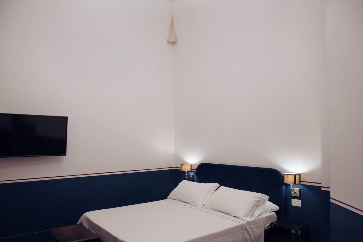 A hotel room with a bed, two bedside lamps, a flat-screen TV mounted on the wall, and a small table.