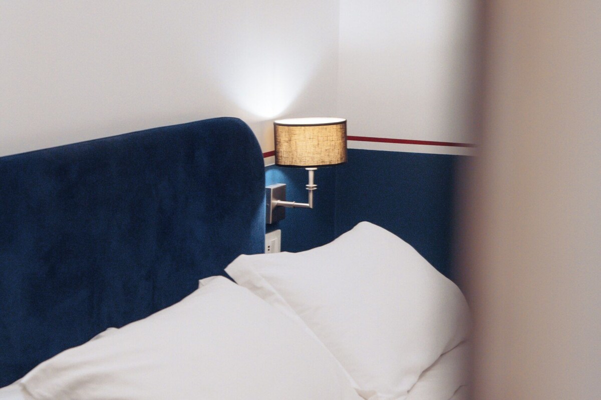 A bed with white bedding and a blue headboard in a room with a wall-mounted lamp.