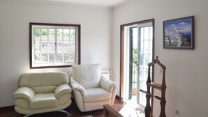 A room with two white armchairs, a wooden chair, and a large window with a view of a building and trees. - Quinta de Maiores (Ovil)