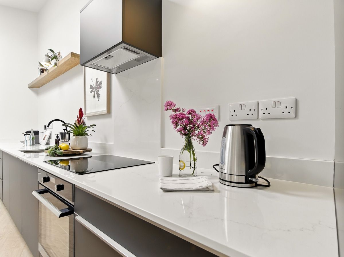 Una cocina moderna con encimera blanca, electrodomésticos de acero inoxidable, una cafetera y un florero con flores.