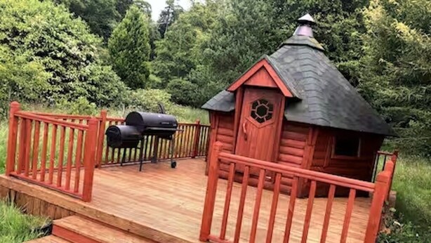 Una cabaña de madera con techo rojo y una chimenea, rodeada por una terraza de madera y barandilla roja.