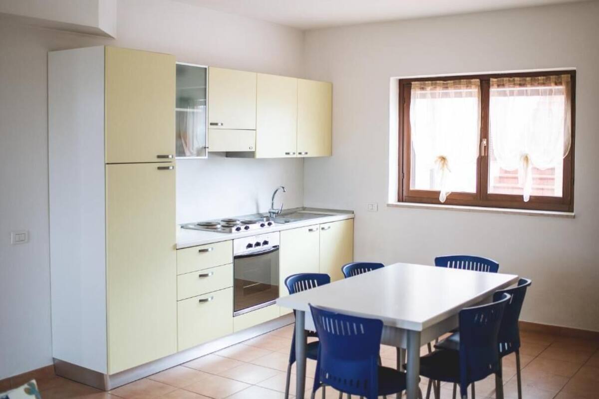 A compact kitchen with a dining area, featuring a white table and blue chairs, a window with curtains, and light-colored cabinets.