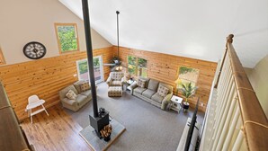 A cozy loft-style living room with wooden walls, a fireplace, and a view of greenery through large windows. - Timber Ridge Retreat by Chatuge Home Concierge | Serene Mountain Escape · Stunning Year-Round Views · Gated Community (Hiawassee)