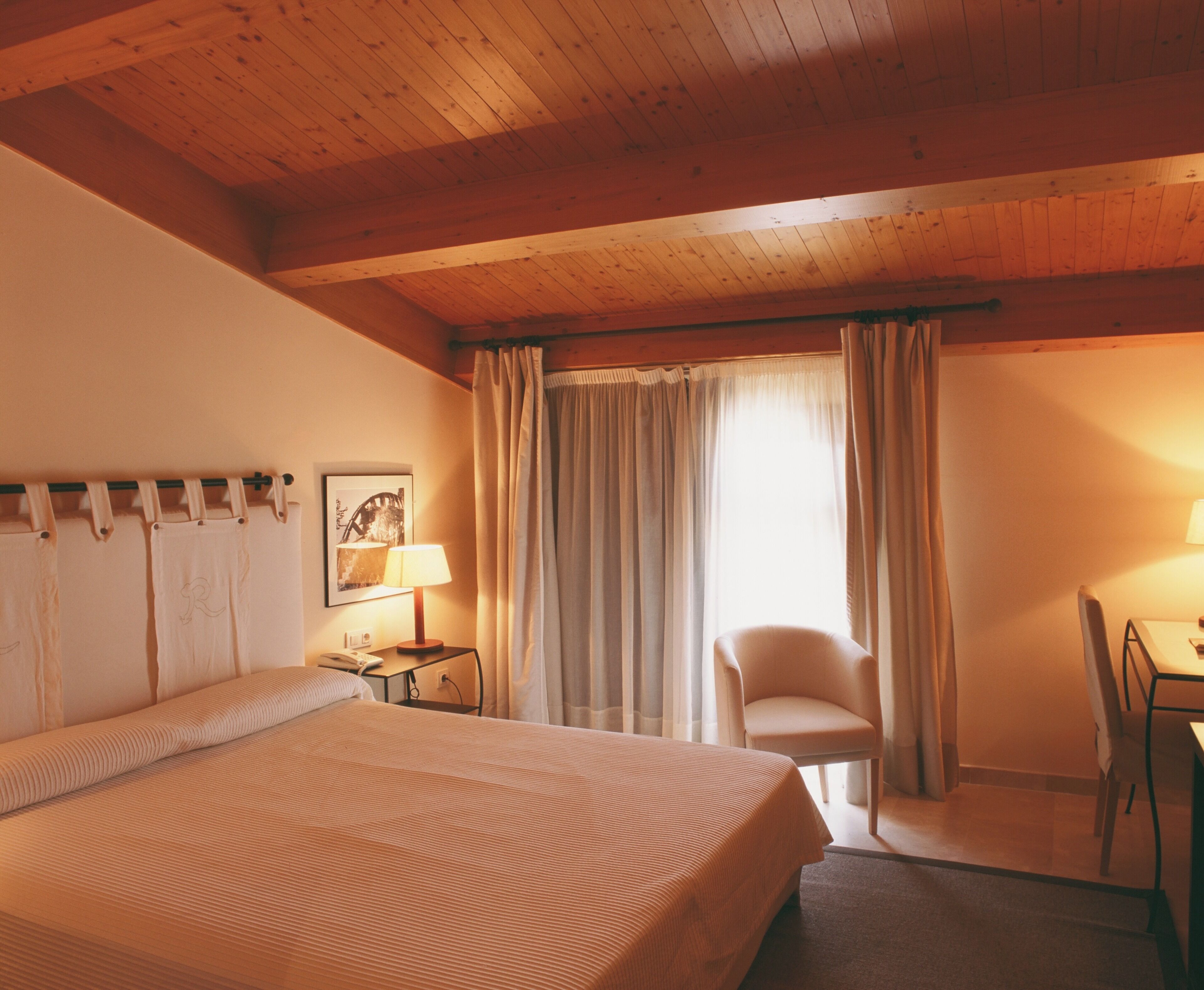 Ein Hotelzimmer mit Holzboden, einem Bett mit gestreifter Bettdecke, einem Schreibtisch mit Lampe, einem Sessel und einem Fenster mit Vorhängen.