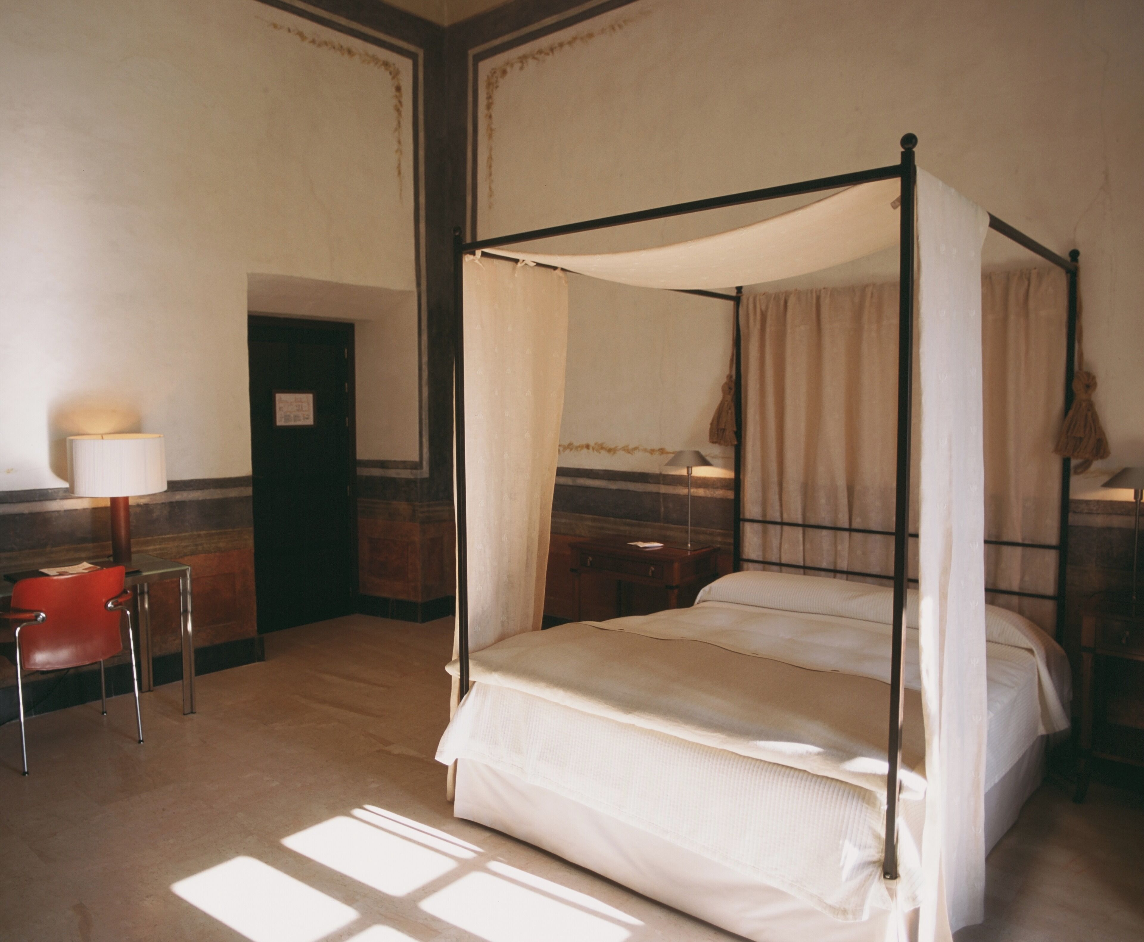 Una cama con dosel y cuatro columnas, una mesita de noche de madera, una silla roja y una lámpara.