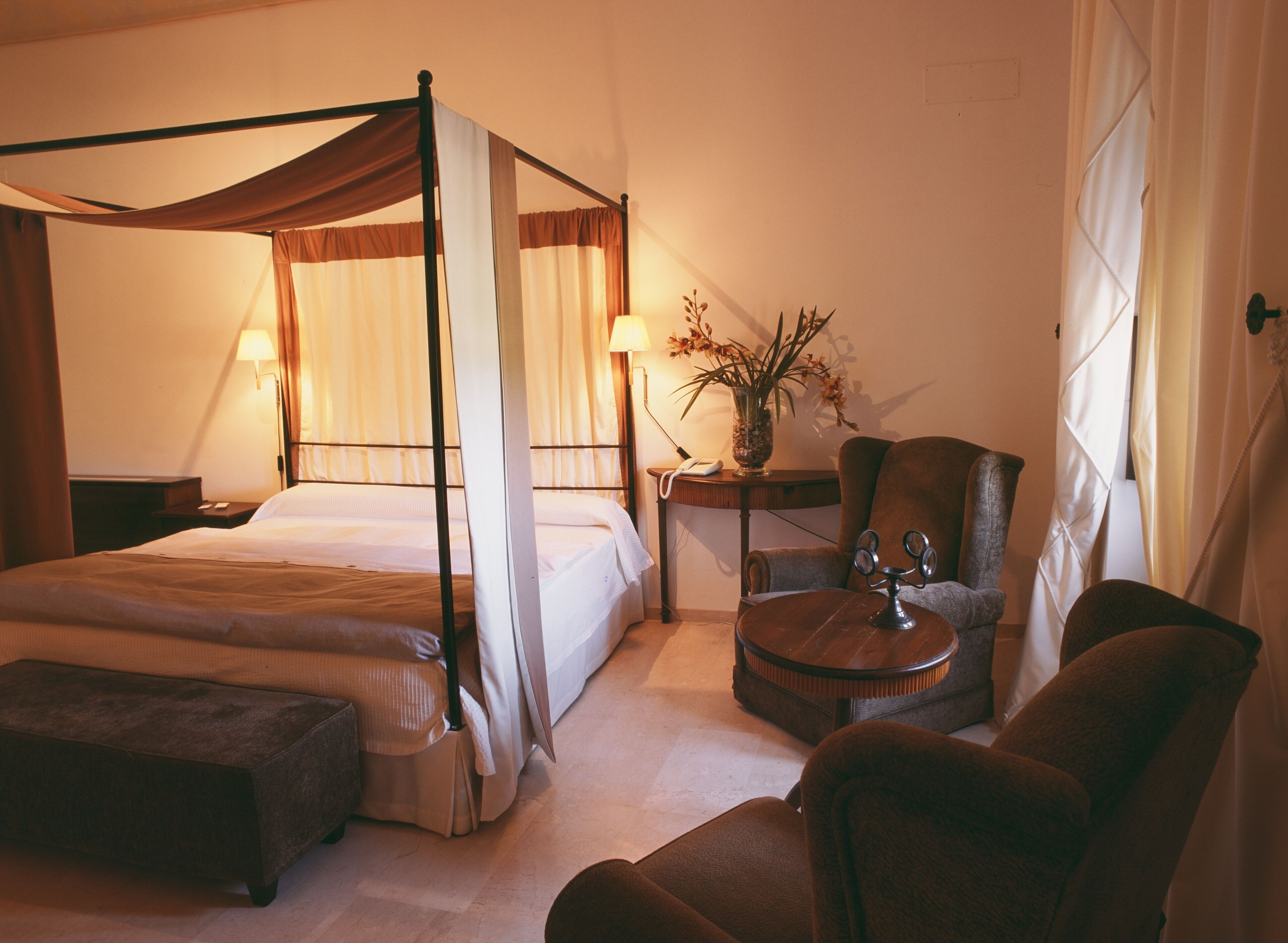 Un lit à baldaquin, une table de chevet avec une lampe, une chaise, une petite table avec un vase et un banc.